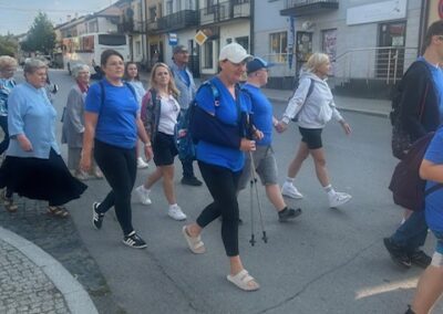 Grupa uczestników idzie ulicą w słoneczny dzień; część osób ma kijki do nordic walking i niebieskie koszulki.