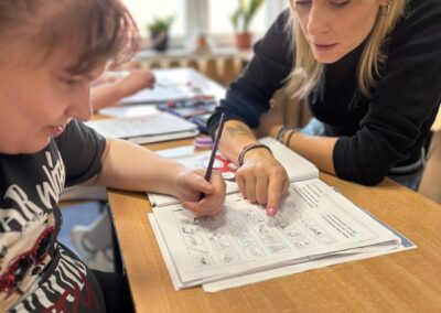 Prowadząca pochyla się nad stołem i wskazuje miejsce na karcie pracy pomagając uczestnikowi wykonać zadanie.