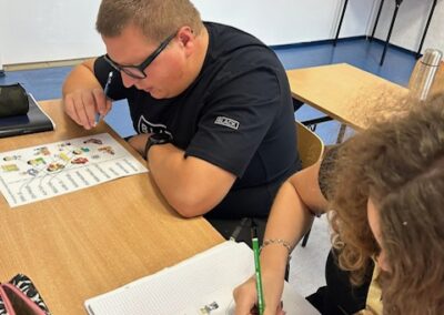 Dwie osoby siedzą przy stole. Jedna przegląda polecenia na karcie a druga zapisuje odpowiedzi na swojej pracy.