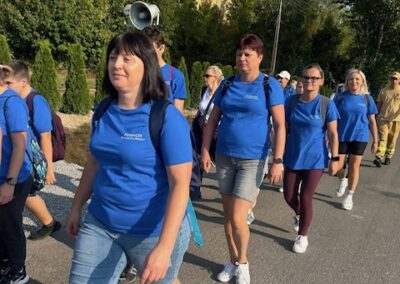 Uczestnicy marszu idą poboczem drogi obok zieleni; widać kolumnę osób w sportowych ubraniach.