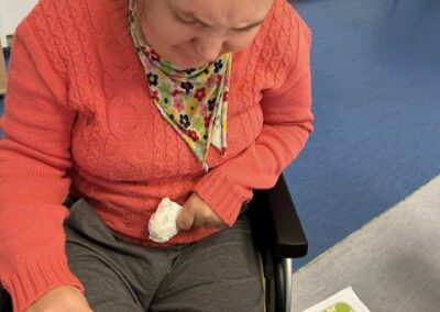 Osoba na wózku wskazuje palcem elementy na ilustrowanej karcie z zadaniami podczas pracy przy stole.