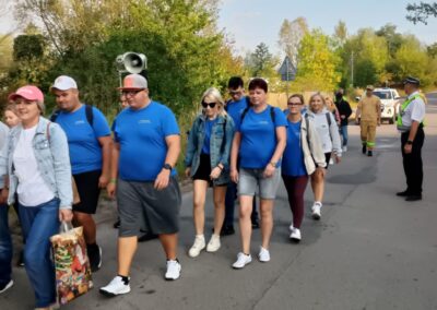 Kolumna uczestników idzie drogą, a obok widać policjanta kierującego ruchem i samochody zatrzymane na czas przejścia.