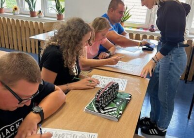 Kilka osób siedzi przy dużym stole i pracuje na kartach pracy a prowadząca stoi obok i udziela wskazówek.