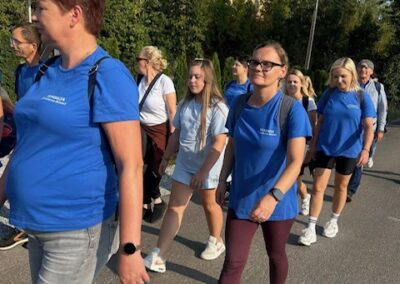 Uczestnicy wydarzenia idą jeden za drugim poboczem; większość ma sportowe ubrania i plecaki.