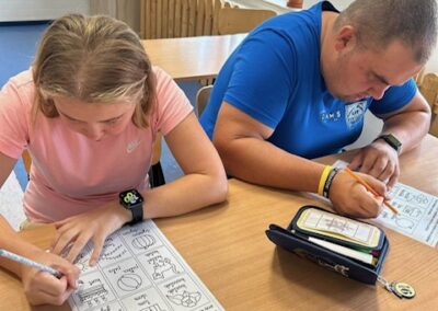 Dwóch uczestników siedzi przy stole i rozwiązuje zadania na wydrukach a na blacie widać piórniki i długopisy.