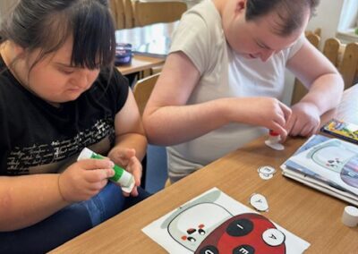 Dwie osoby przy stole przyklejają papierowe kółka na ilustracjach biedronek korzystając z kleju w sztyfcie.