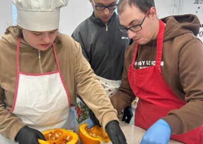Trzy osoby, ubrane w fartuchy i rękawiczki, pracują razem nad wydrążaniem nasion z przekrojonych połówek dyni. Na stole leży papier do pieczenia z zebranymi nasionami.