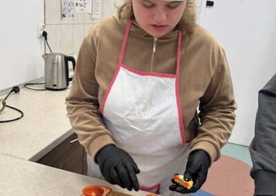 Młoda dziewczyna w fartuchu i czapce kucharskiej, w czarnych rękawiczkach, starannie wyjmuje nasiona z przekrojonej na pół czerwonej papryki leżącej na drewnianej desce do krojenia.