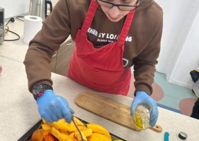 Młody mężczyzna w czerwonym fartuchu i niebieskich rękawiczkach polewa oliwą kawałki dyni i papryki na blasze do pieczenia, używając łyżki, by równomiernie rozprowadzić tłuszcz.