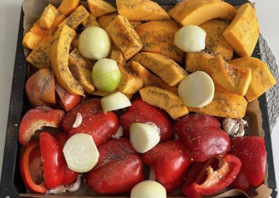 Bliska fotografia tacy do pieczenia wypełnionej kawałkami dyni, papryki, czosnku i ćwiartkami cebuli, obficie posypanych ziołami i solą. Warzywa mają intensywne kolory pomarańczowy i czerwony.