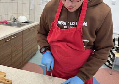 Bliski kadr na młodego mężczyznę w czerwonym fartuchu i niebieskich rękawiczkach, który starannie kroi bagietkę na równe kromki na małej, niebieskiej desce do krojenia.