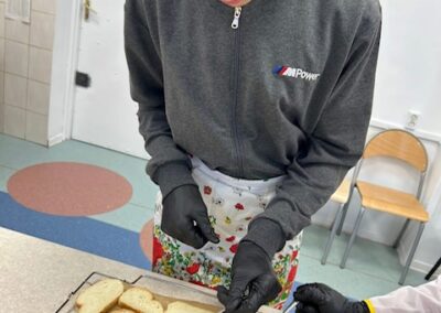 Młody mężczyzna w okularach, szarej bluzie i fartuchu, w czarnych rękawiczkach, delikatnie nakłada oliwę ziołową łyżeczką na kromki bagietki ułożone na tacy do pieczenia.