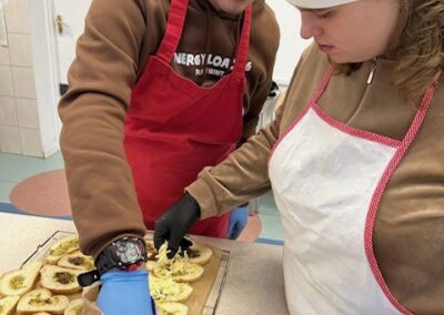 Dwójka kucharzy w fartuchach i rękawiczkach posypuje przygotowane kromki chleba ziołowego startym serem z torebki. Widoczne jest skupienie na dokładnym rozprowadzeniu sera.