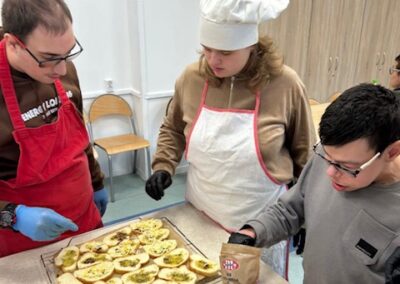 Trzy osoby stoją przy stole, nad tacą gotowych do pieczenia grzanek z oliwą i serem. Mężczyzna z prawej, w szarej bluzie, trzyma torebkę z serem, a pozostali nadzorują.