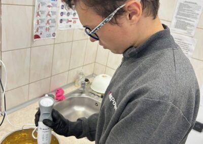 Młody mężczyzna w okularach i szarej bluzie, w czarnych rękawiczkach, używa blendera ręcznego do miksowania upieczonej dyni i papryki w metalowej misce, tworząc gęstą zupę lub pastę.