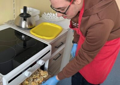 Młody mężczyzna w okularach, fartuchu i niebieskich rękawiczkach wkłada blachę z upieczonymi, złocistymi grzankami do otwartego piekarnika kuchennego, stojącego w jasnej kuchni.