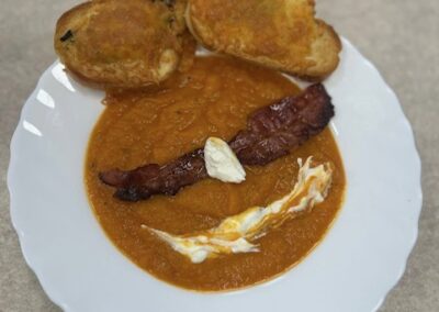 Zbliżenie na danie: kremowa, pomarańczowa zupa na białym talerzu, udekorowana plasterkiem bekonu i kleksami śmietany. Obok zupy leżą dwie zapieczone grzanki.