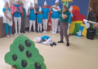 Grupa osób w kostiumach Smerfów podczas przedstawienia teatralnego z kolorową dekoracją.