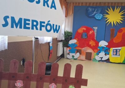 Dekoracja Wioski Smerfów z papierowymi postaciami, domkami, tablicą i płotem z kwiatami.
