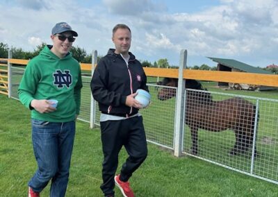 Dwaj mężczyźni idą obok ogrodzenia, za którym stoi kucyk na trawiastej łące.