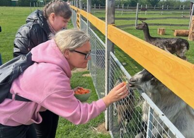 Dwie kobiety karmią kozę przez ogrodzenie na zielonym pastwisku pod niebieskim niebem.