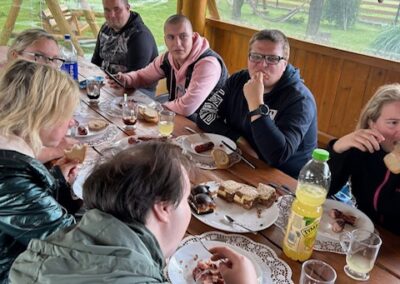 Grupa siedzi przy drewnianym stole na zewnątrz, jedząc przekąski i pijąc napoje.