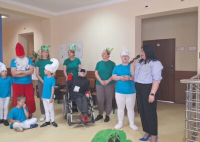 Grupa osób w kolorowych kostiumach, w tym osoba na wózku, jedna przemawia przez mikrofon.