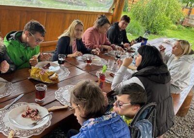 Grupa osób spożywa posiłek przy drewnianym stole pod zadaszeniem na zewnątrz.