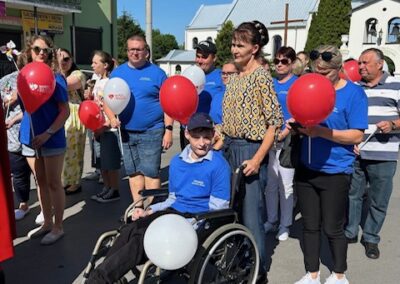 Grupa osób z balonami na ulicy, wśród nich chłopiec na wózku, w słoneczny dzień.