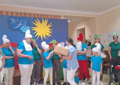 Grupa osób w kostiumach postaci z bajki na tle dekoracji słonecznej, wręczanie prezentu.