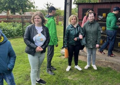 Grupa osób stoi na trawie obok drewnianej budki z klatkami na tle drzew i ogrodzenia.