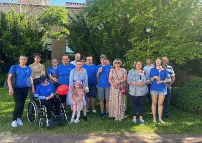 Grupa osób na trawniku przed budynkiem, niektórzy w niebieskich koszulkach, dzieci z czerwonym balonem.