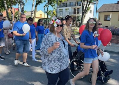Grupa osób idzie ulicą w słoneczny dzień, niesie czerwone i białe balony oraz flagi.