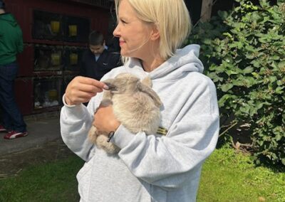 Kobieta w jasnoszarej bluzie trzyma króliczka na tle zielonej trawy i krzewów.