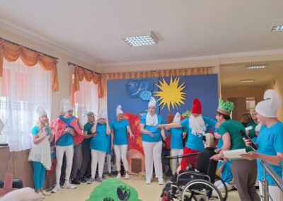 Grupa osób w kostiumach smerfów podczas występu w sali z dekoracjami słonecznymi i chmurnymi.