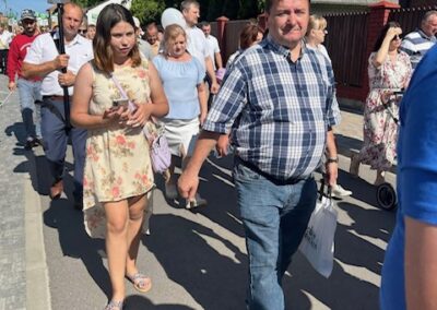 Grupa ludzi maszeruje ulicą w słoneczny dzień, w tle budynki i zieleń.