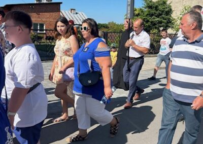 Ludzie maszerują ulicą w słoneczny dzień, w tle budynki i głośniki na słupie.