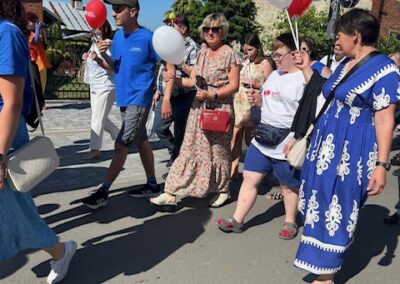 Ludzie idą ulicą w słoneczny dzień, niosąc białe i czerwone balony z napisem.