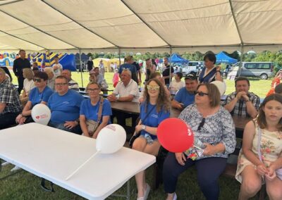 Osoby siedzą pod namiotem na ławkach podczas plenerowego wydarzenia z balonami i atrakcjami w tle.