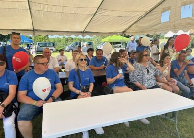 Ludzie siedzą przy stołach pod namiotem na zewnątrz, niektórzy z balonami.