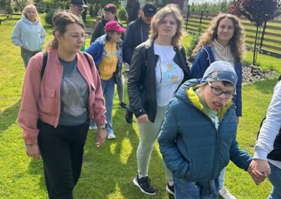 Grupa ludzi spaceruje po trawniku w ogrodzie, kilka osób idzie za chłopcem trzymającym rękę.