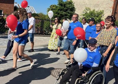 Ludzie idą ulicą z czerwonymi i białymi balonami, osoba na wózku, słoneczna pogoda.
