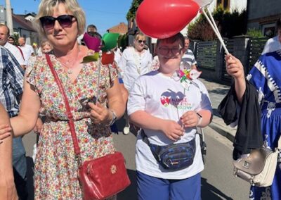 Grupa osób idzie ulicą w słoneczny dzień, kobieta w kwiecistej sukience i osoba z balonami.