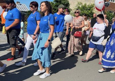 Ludzie idą ulicą w słoneczny dzień, część w niebieskich koszulkach, niektórzy z balonami.