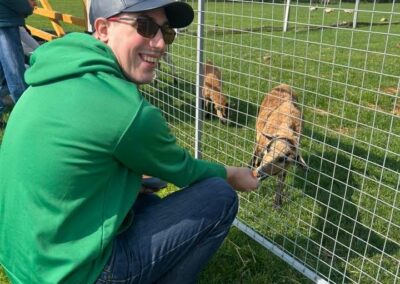 Mężczyzna w zielonej bluzie karmi zwierzęta przez metalowe ogrodzenie na trawniku.