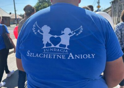 Mężczyzna w niebieskiej koszulce z logo Fundacji Szlachetne Anioły stoi na ulicy w grupie ludzi.