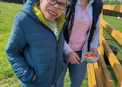 Mężczyzna w niebieskiej kurtce i czapce stoi obok kobiety w różowej bluzce z miską marchewek przy drewnianym płocie.