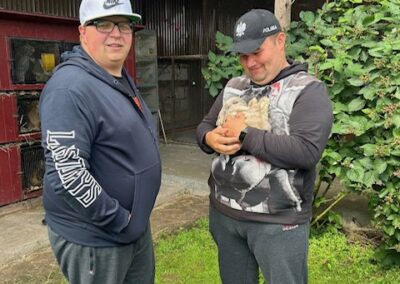 Dwóch mężczyzn stoi na trawie przed budynkiem gospodarczym, jeden trzyma pisklęta.