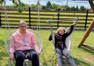 Dwoje młodych ludzi siedzących na huśtawkach ogrodowych na trawiastej działce przy drewnianym płocie.