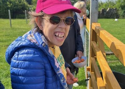 Osoba w różowej czapce i niebieskiej kurtce stoi przy drewnianym ogrodzeniu na zielonej posesji.